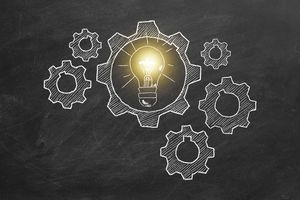 A chalkboard-style illustration featuring several white outlined gears interconnected in a mechanical arrangement. At the center of the composition is a large gear containing a glowing yellow light bulb, symbolizing an idea or innovation. The surrounding gears are drawn in white chalk, creating a sense of movement and collaboration, while the light bulb stands out as the focal point with its bright illumination. The background is a dark, textured chalkboard surface, reinforcing the concept of brainstorming, creativity, and problem-solving.