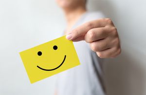 A close-up image of a person holding a bright yellow rectangular card with a simple black smiley face printed on it. The card is positioned prominently in the foreground, while the person’s torso and hand are partially visible in the background, slightly out of focus. The background is plain and light-colored, creating a clean and minimalistic setting that emphasizes the cheerful design on the card. The overall composition conveys positivity, friendliness, and encouragement.