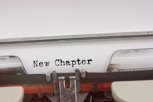 A close-up view of an old-fashioned typewriter with a sheet of white paper inserted. The paper displays the words ‘New Chapter’ typed in black, slightly distressed typewriter-style font. The metal type guide and ruler are visible at the top, and the typewriter’s mechanical parts, including the ribbon and keys, are partially shown at the bottom. The overall composition emphasizes the typed phrase, symbolizing beginnings, transitions, or fresh starts.'