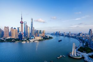 A panoramic view of Shanghai’s modern skyline along the Huangpu River under a clear blue sky. The foreground shows the river curving through the city, with boats visible on the water. On the left side of the image, the iconic Oriental Pearl Tower stands out with its spherical design, alongside other tall skyscrapers including the Shanghai Tower and Jin Mao Tower. The right side of the image features a white triangular monument near the riverbank and a stretch of urban buildings extending into the distance. The scene is brightly lit by daylight, highlighting the contrast between the glass skyscrapers and the calm river.