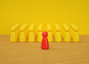 The image shows a single red game pawn standing at the front center of the frame on a light wooden surface, facing a group of yellow game pawns arranged in several neat rows behind it against a solid bright yellow background.