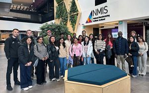 A group of MBA participants standing together in the atrium of the National Manufacturing Institute Scotland (NMIS). The modern interior features a large NMIS sign, living green walls, contemporary architecture, and a blue upholstered bench in the foreground.